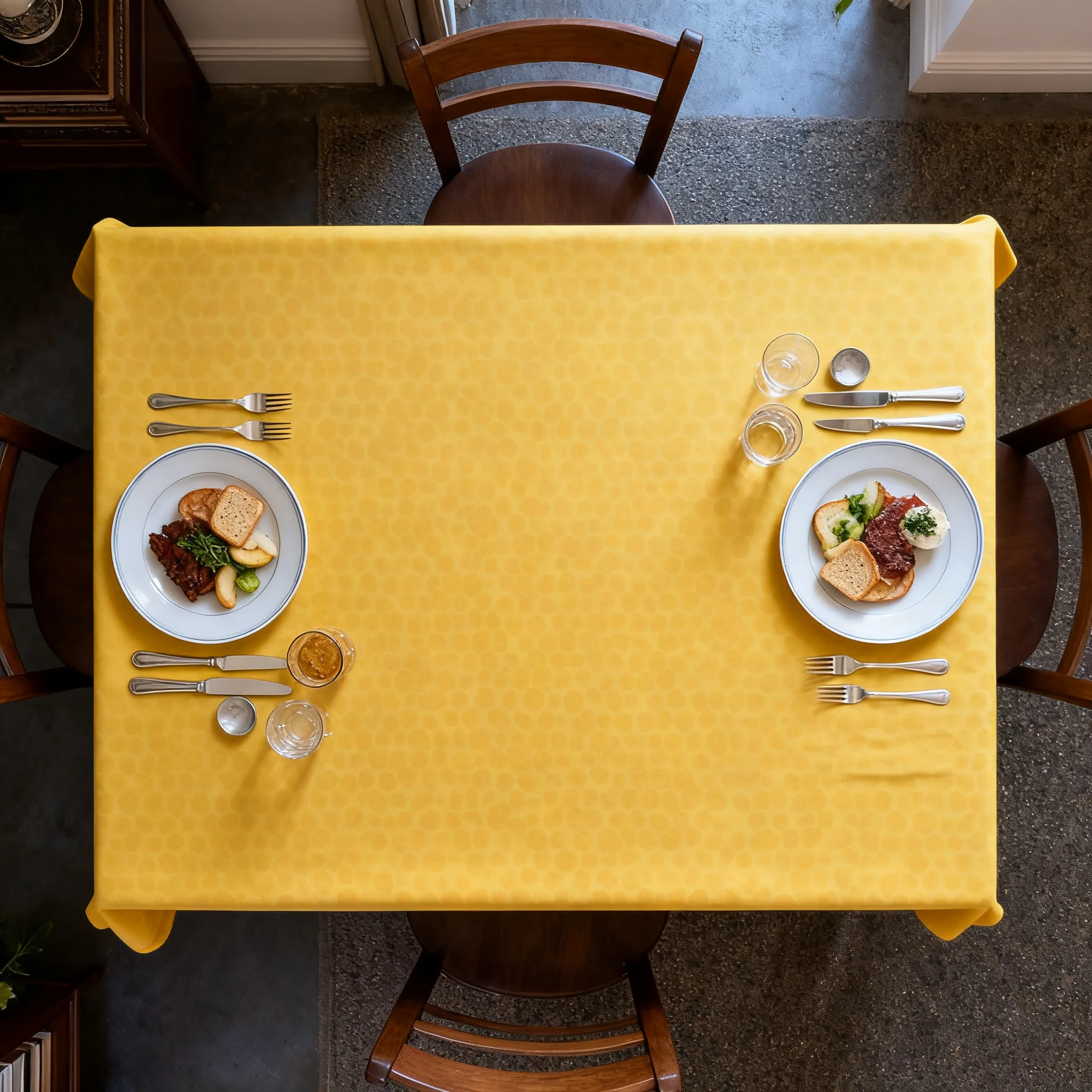 Overhead view of a yellow tablecloth set for two with plates, cutlery, and glasses at a wooden table.