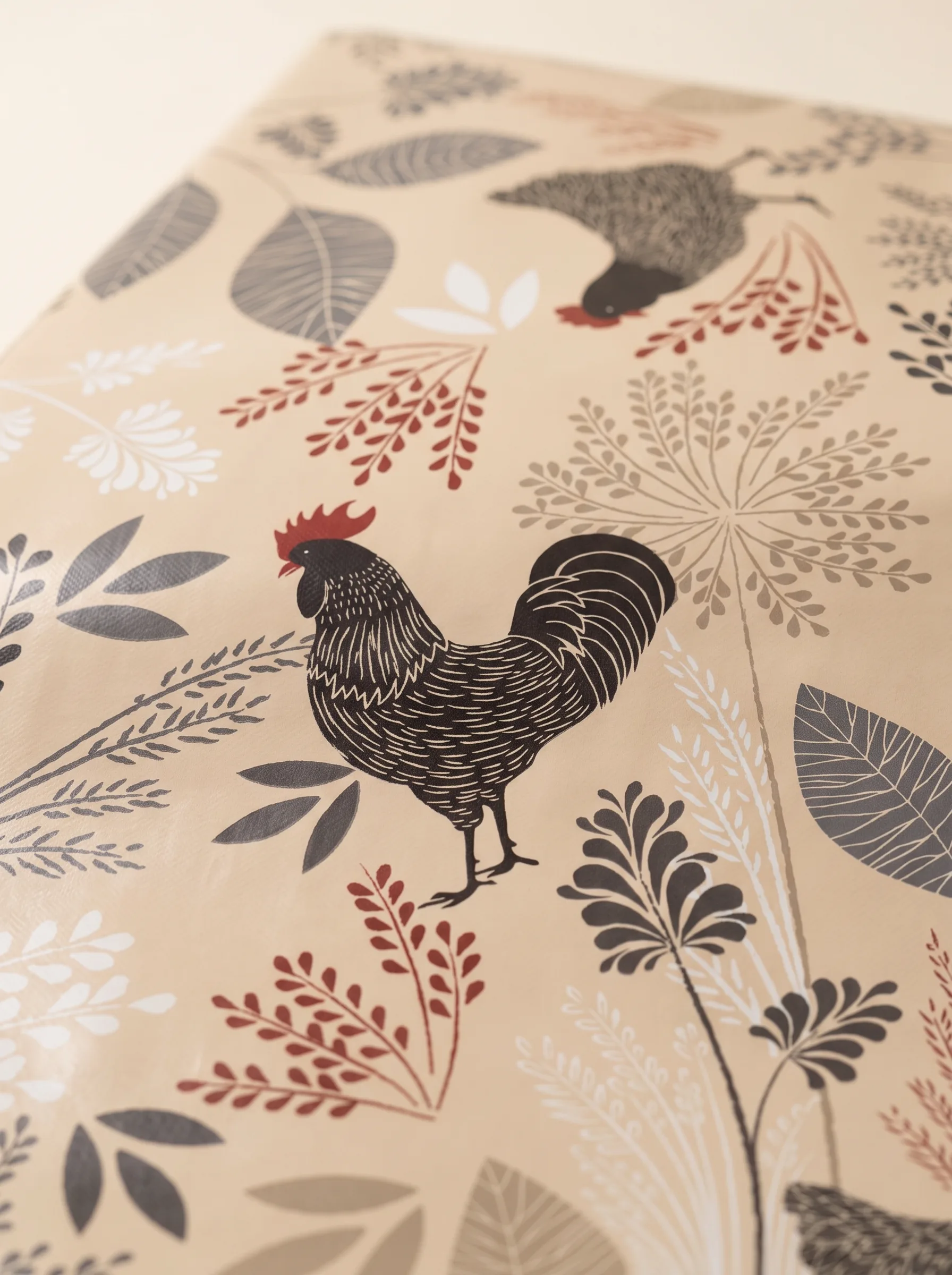 Gros plan sur le motif de la toile cirée déco Cotcot beige – coq dessiné à l'encre fine, crête rouge vif, feuillages graphiques gris anthracite et rouge bordeaux sur fond beige sable