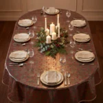 Toile cirée transparente Étoile Rouge sur grande table ovale dressée pour dix vue du dessus – étoiles rouges all-over, biais beige, serviettes ivoire, verres à vin, centre de table végétal houx et eucalyptus avec bougies piliers blanches, parquet en chêne visible sous la toile, lustre en fer forgé